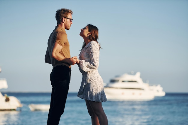 背景の海とヨット幸せな若いカップルは彼らの休暇に一緒に晴れた日中の屋外
