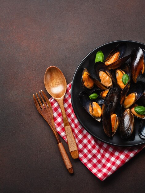 Seafood mussels and basil leaves in a black plate with towel, wooden spoon and fork on rusty background. Top view.