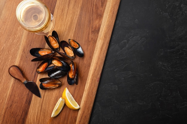 Seafood mussels and basil leaves in a black plate with wineglass, lemon, knife on wooden board and stone table. Top view with place for your text.