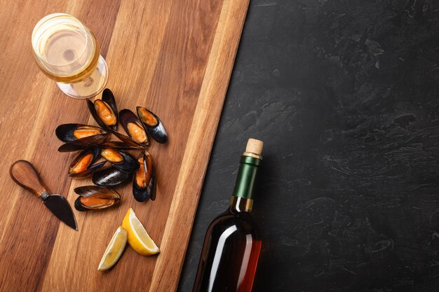 Seafood mussels and basil leaves in a black plate with wineglass, lemon, knife on wooden board and stone table. Top view with place for your text.