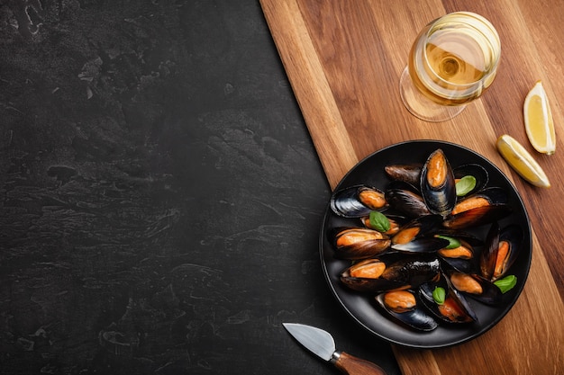 Seafood mussels and basil leaves in a black plate with wineglass, lemon, knife on wooden board and stone table. Top view with place for your text.