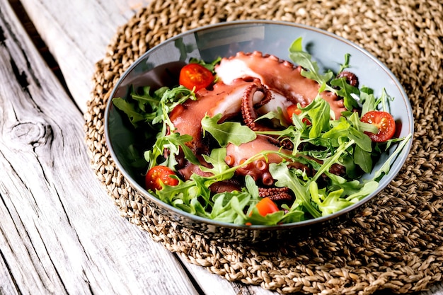 Seafood salad. Coocked tentacles of octopus on blue ceramic plate served with rocket leaf aragula and cherry tomato salad over grey wooden surface and wicker lining.