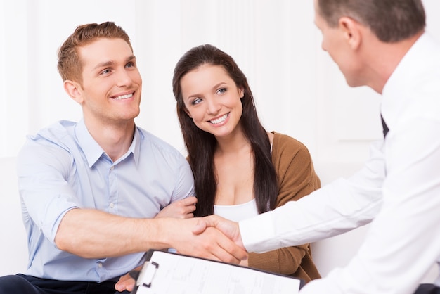 Sealing a deal. Happy young man shaking hand to financial agent while sitting together with his wife