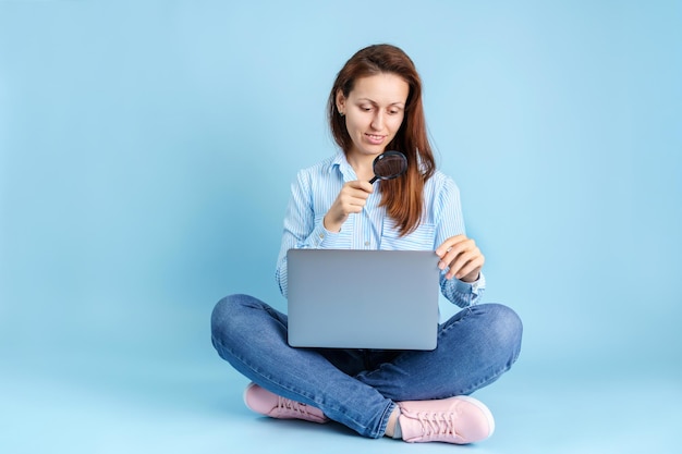 Search for information on the Internet. A young adult girl is sitting with a laptop on her lap and holding a magnifying glass in her hands on a blue background