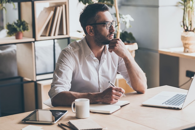 In search of inspiration. Thoughtful mature man holding pen and looking away while sitting at his working place in office