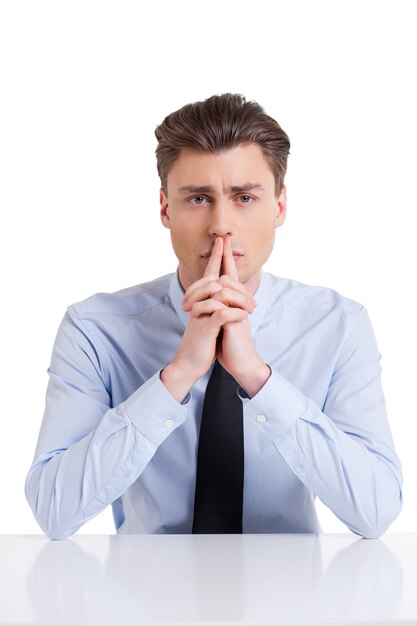 In search of solution. Thoughtful young man in shirt and tie holding fingers on mouth and looking at camera while sitting at the table and isolated on white