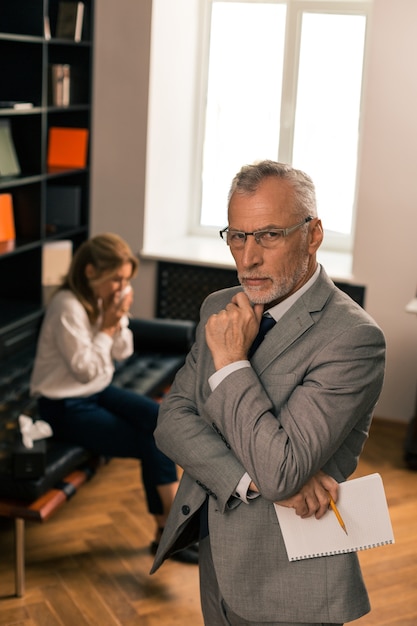 Searching for answers. Serious psychologist standing in his office and thinking next to his crying female patient