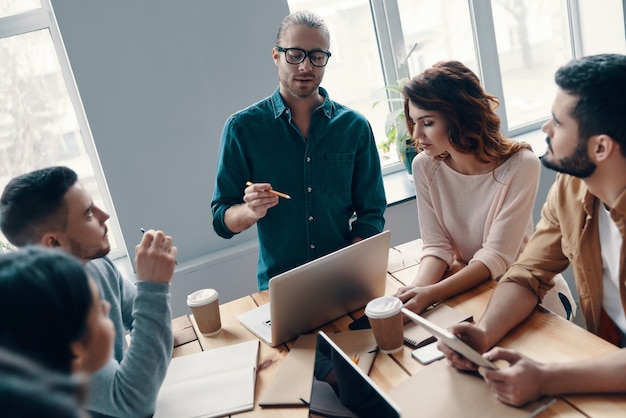 Searching for right decision. Top view of young modern people in smart casual wear discussing something while working in the creative office