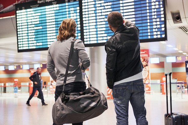 Searching their plane and when it arrives. Photo of two comrades situating in airport near flight information display system