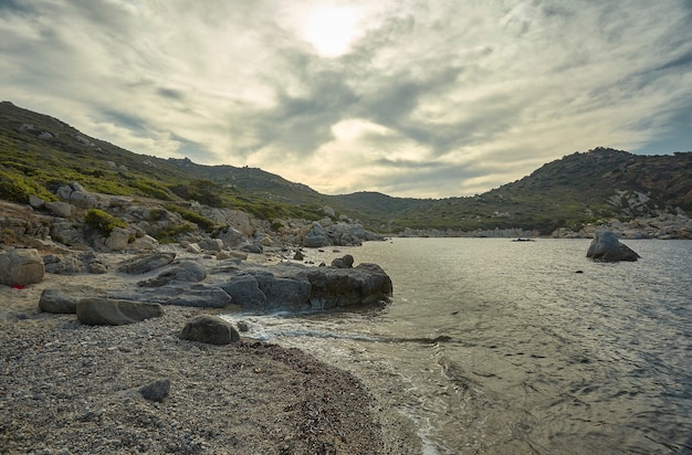 Seascape al tramonto content una magnifica spiaggia mediterranea con i promontori rocciosi e le montagne sotto un cielo nuvoloso con spiragli di sole.