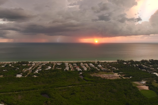 Seashore town with dark houses and tall lighthouse on ocean\
shore with blinking light at stormy night for vessels navigation\
thunderstorm with lightnings over sea water posing danger for\
ships