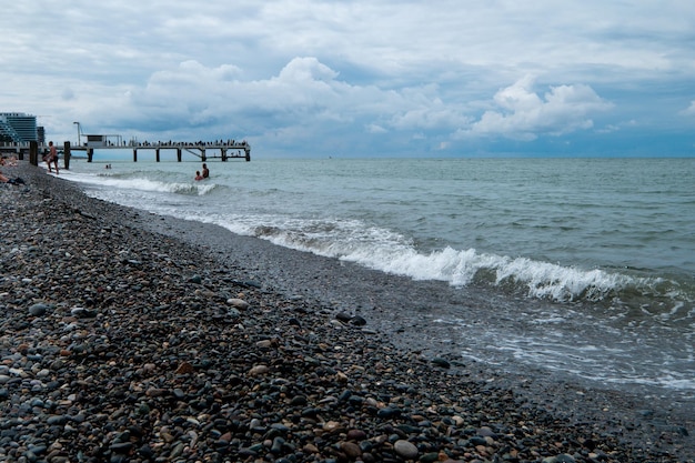 Photo seashore with waves and pebbles and a pier in the distance with vacationing people