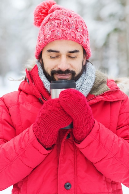 season, christmas, drinks and people concept - happy smiling young man with thermo cup drinking hot tea in winter forest