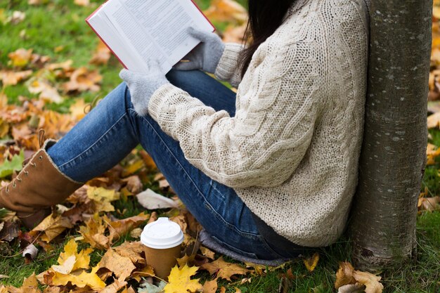 Фото Сезон, литература, образование и концепция людей - крупный план молодой женщины, читающей книгу и пьющей кофе из бумажного стаканчика в осеннем парке