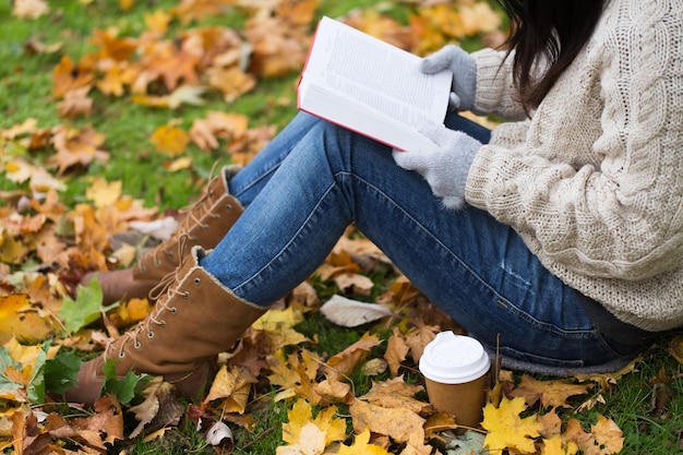 season, literature, education and people concept - close up of young woman reading book and drinking coffee from paper cup in autumn park