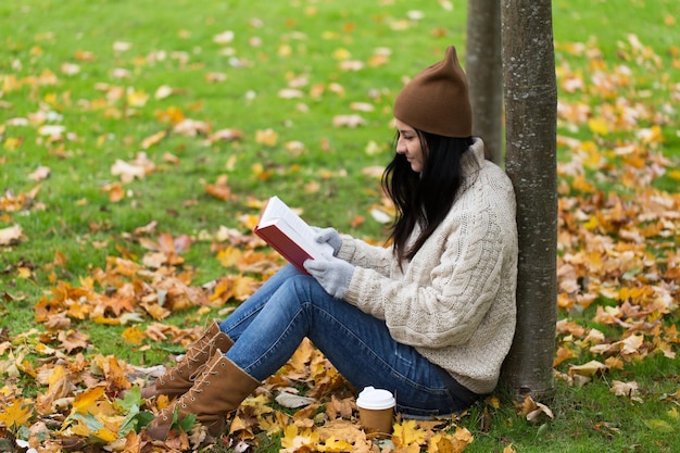 season, literature, education and people concept - young woman reading book and drinking coffee from paper cup in autumn park