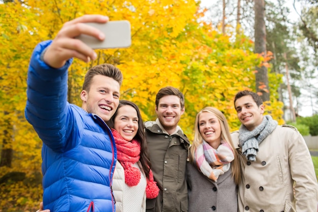 сезон, люди, технологии и концепция дружбы - группа улыбающихся друзей со смартфоном, делающих селфи в осеннем парке
