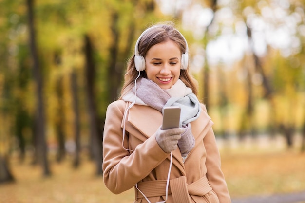 season, technology and people concept - beautiful happy young woman with headphones listening to music on smartphone walking in autumn park