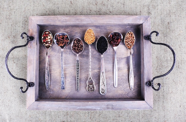 Seasonings in metal spoons on wooden tray on fabric background