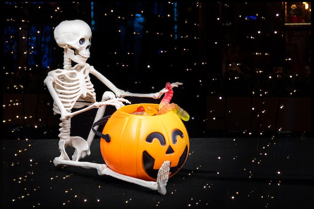 A seated skeleton pulls out of a bucket in the shape of a pumpkin candy on a black background with a starry bokeh