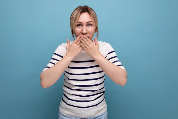 Secret blond girl covered her mouth with her hands on a blue background with copyspace