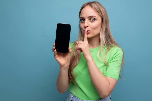 Секретная яркая блондинка в повседневной одежде, держащая смартфон вертикально с мокапом вперед на синем