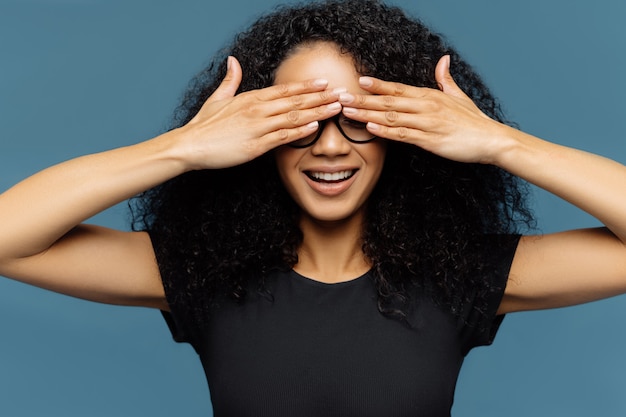 Secret woman with curly hair, covers eyes, wears spectacles, waits for surprise, dressed in black casual t shirt