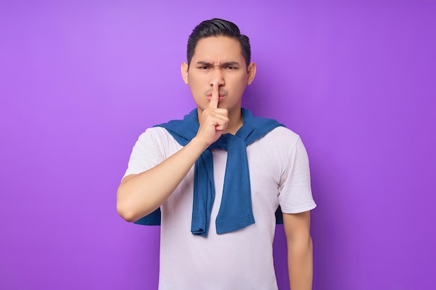 Secret young Asian man wearing white tshirt say hush be quiet with finger on lips shhh gesture isolated over purple background people lifestyle concept