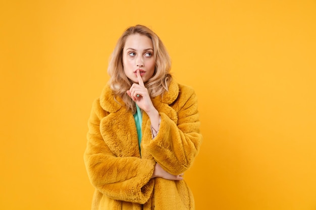 Secret young blonde woman in yellow fur coat posing isolated on orange background studio portrait. People lifestyle concept. Mock up copy space. Saying hush be quiet with finger on lips shhh gesture.