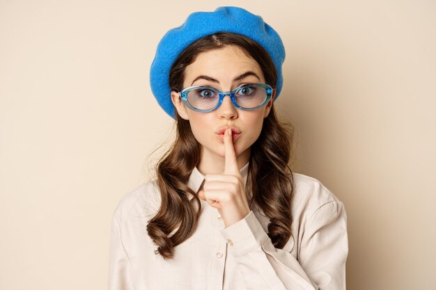 Secret young cute woman in sunglasses shushing showing taboo hush sign press finger to lips standing over beige background person