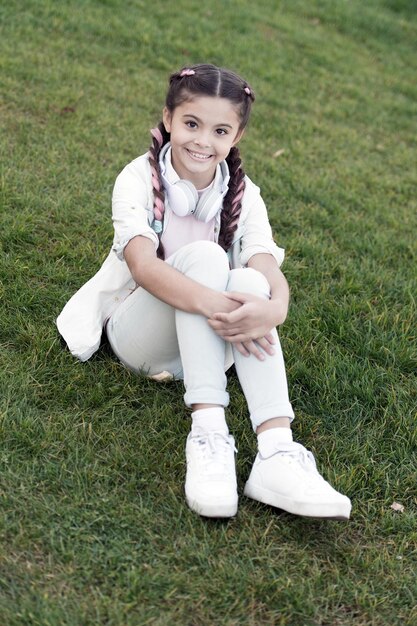 Secrets to raising happy child Girl cute kid green grass background Healthy emotional happy kid relaxing outdoors Get happy yourself Girl braids hairstyle and modern headphones enjoy relax