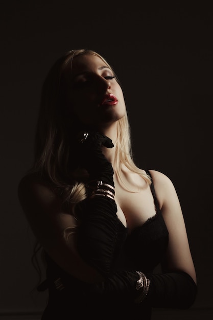 Seductive blonde woman in a black lace lingerie and silk gloves posing in the shadows