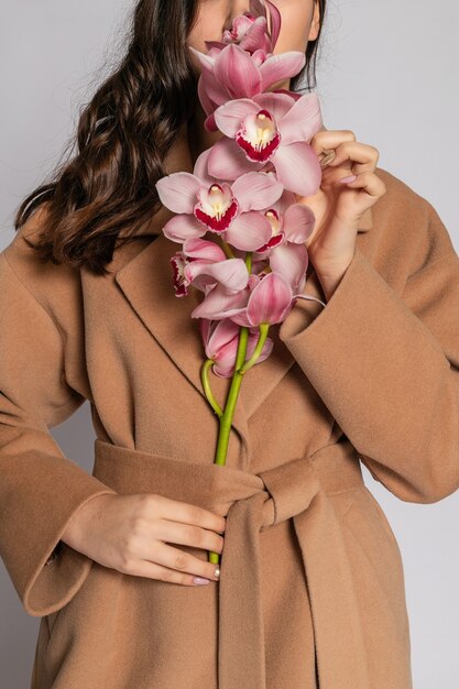 Seductive blonde woman in brown jacket posing in studio on grey background. Fashion portrait of elegant model in pastel casual spring outfit. Beautiful girl with healthy skin and branch orchid