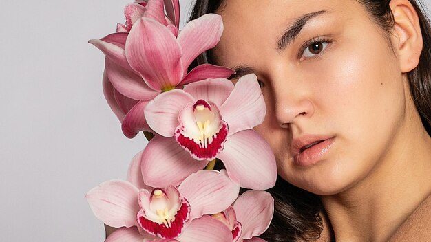 Foto donna bionda seducente con giacca marrone che posa in studio su sfondo grigiomodello elegante in abito primaverile casuale pastellobella ragazza con pelle sana e rami di orchidee 169 formato panoramico