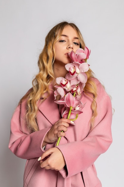 Соблазнительная блондинка в розовой куртке позирует в студии на сером фоне. Фасонный портрет элегантной модели в пастельном повседневном весеннем наряде. Красивая девушка со здоровой кожей и веткой орхидеи
