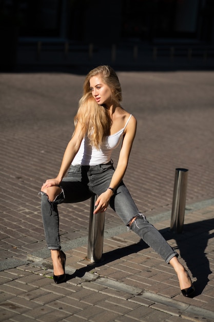 Соблазнительная блондинка в повседневных рваных джинсах и футболке позирует у парковочного ограждения