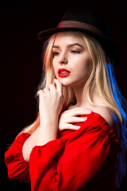 Соблазнительная блондинка в красной блузке и шляпе позирует в тени с синим и красным светом