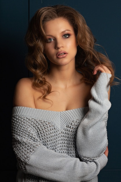 Seductive blonde woman with curly hair wearing knitted sweater with naked shoulders