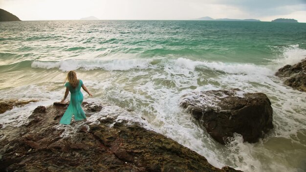 魅惑的な長髪の女性は海礁の岩の上を歩く石嵐の曇り海青い水着ドレスチュニックの女性コンセプトは海の熱帯リゾート海岸線旅行観光夏休みで休む