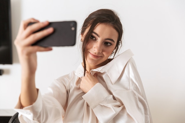 朝のキッチンに立っている間、黒いスマートフォンでselfie写真を撮るシルクの服を着ている魅惑的な女性