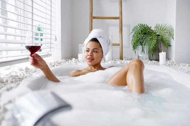 와인 한 잔을 들고 있는 매혹적인 여성은 거품 목욕에서 휴식을 취합니다. 욕조에 있는 여성, 스파의 미용 및 건강 관리, 욕실의 웰빙 트리트먼트, 배경에 자갈 및 촛불