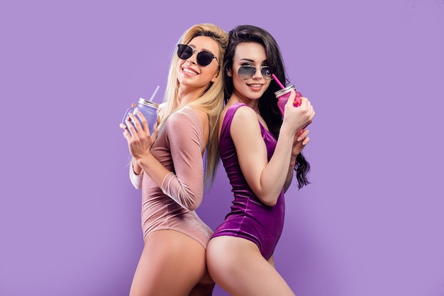 Seductive women in bodysuits standing together with colorful drinking jars on purple wall.