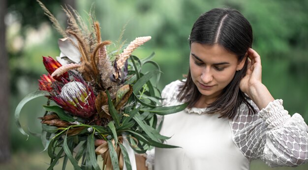 ぼやけた背景に森の花の花束と白いドレスを着た魅惑的な若いブルネットの女性、