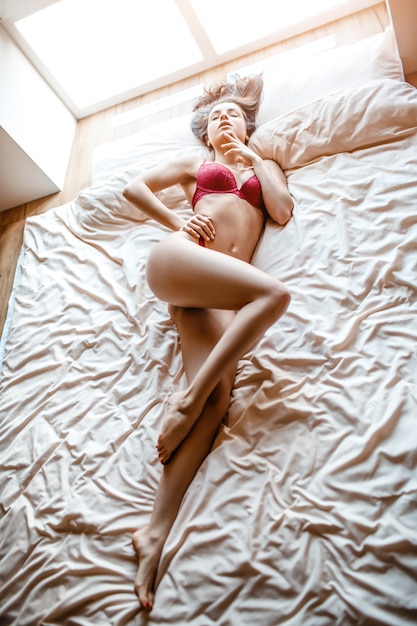 아침에 침대에 매혹적인 젊은 벗은 검은 머리 여자. 누워 즐기는 관능적인 섹시 모델의 세로 사진입니다. 아름다운 빨간 란제리
