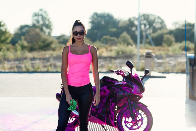 Seductive young woman in pink tshirt poses near sport motorcycle at self service car wash in the