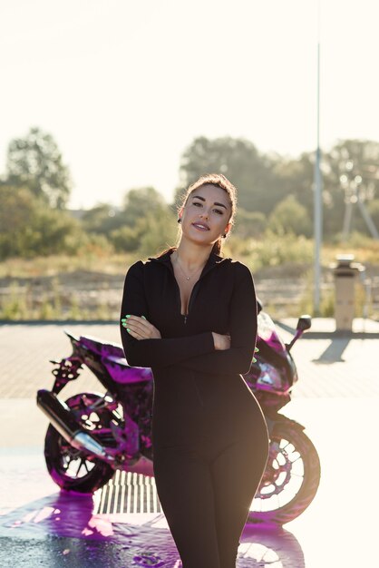 Seductive young woman in tight fitting black suit poses near sport motorcycle at self service car