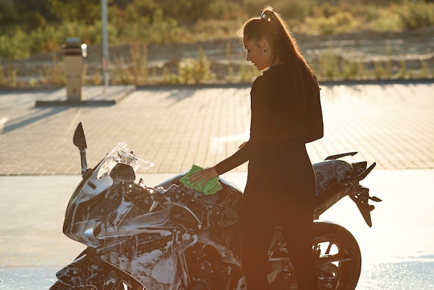 Giovane donna seducente che lava la motocicletta sportiva alla moda e la asciuga dalla schiuma viola. prendersi cura del veicolo.