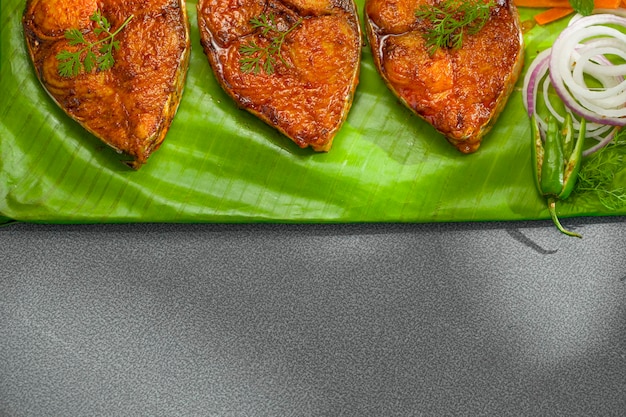 Seer fish fry arranged beautifully and garnished with onion lemon and tomato slices on banana leaf covered base which is placed on solid green texture with abstract shadows on background