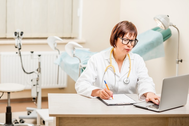 Seinor doctor working with laptop and documents sitting in the gynecological office with gynecological chair on the background