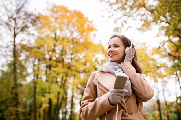 seizoen, technologie en mensenconcept - mooie gelukkige jonge vrouw die met een koptelefoon naar muziek luistert op een smartphone die in het herfstpark loopt